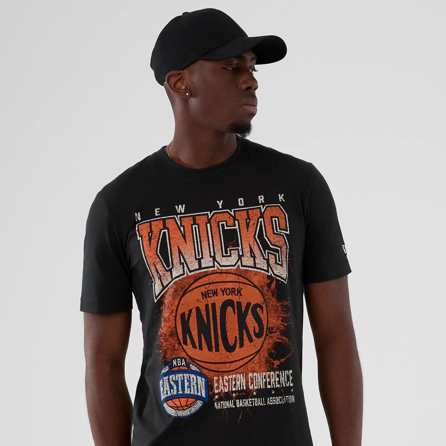 New Era New York Knicks NBA Hardwood Classic Black T-Shirt, front view showing a large basketball graphic with New York Knicks lettering across the chest and an NBA Eastern Conference logo below, worn by a model with a plain black cap.