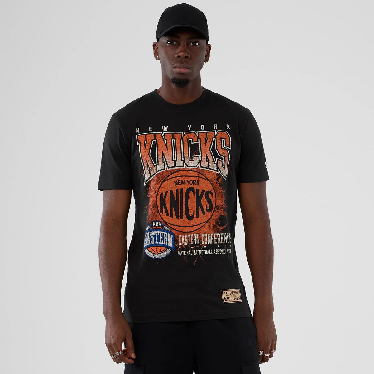 New Era New York Knicks NBA Hardwood Classic Black T-Shirt, front view on-model showing a vintage basketball graphic with New York Knicks lettering in orange and white across the chest and an embroidered New Era logo on the sleeve.