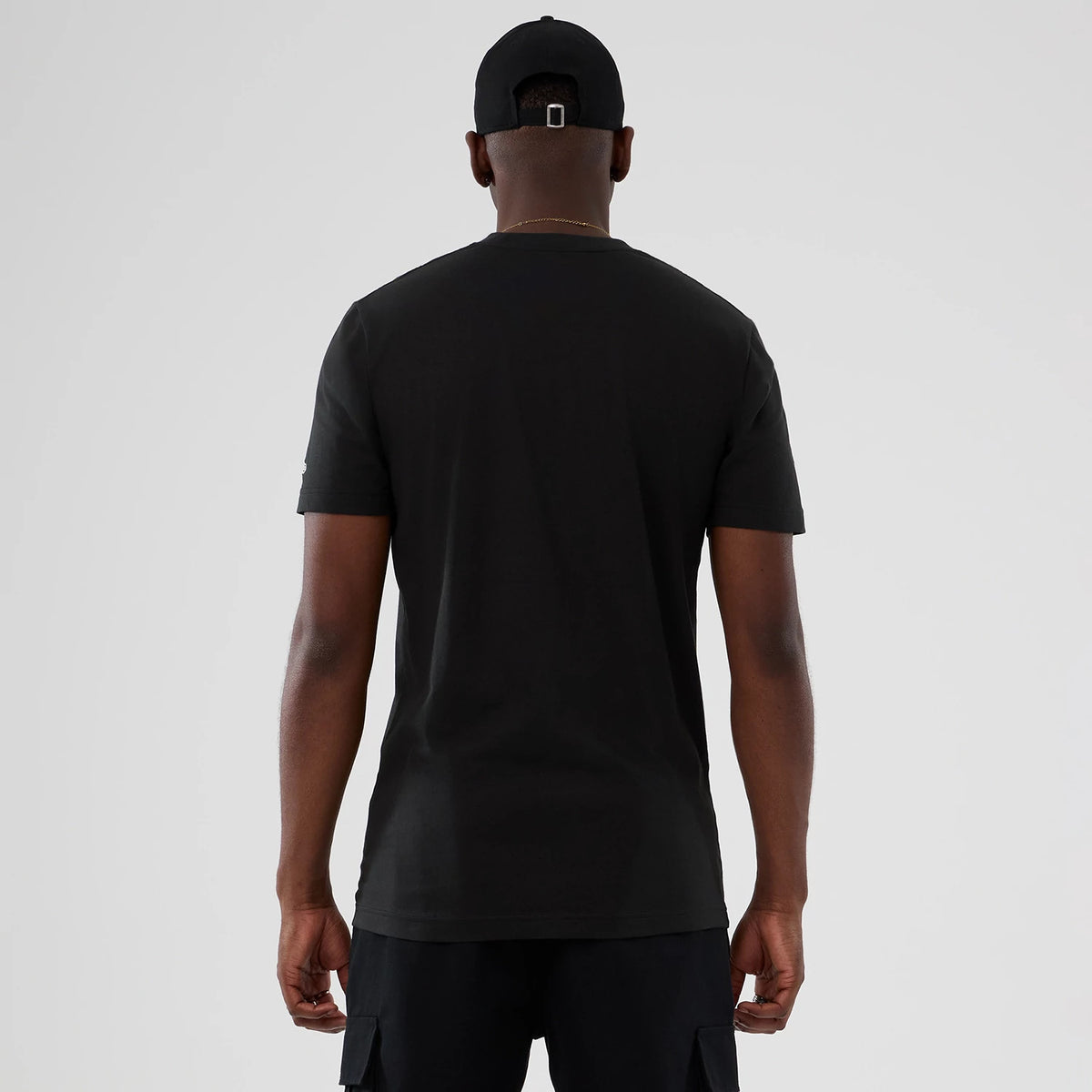 New Era New York Knicks NBA Hardwood Classic Black T-Shirt, back view on a model wearing a matching black cap, showing a plain black back and short sleeves with a small embroidered New Era logo on the left sleeve.