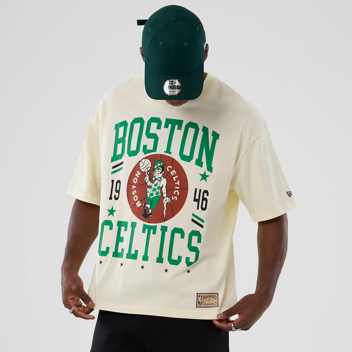 New Era Boston Celtics NBA Hardwood Classic Cream T-Shirt, front view on-model showing large green Boston Celtics lettering and a mascot graphic centred on the chest, with a small woven New Era logo on the left sleeve.