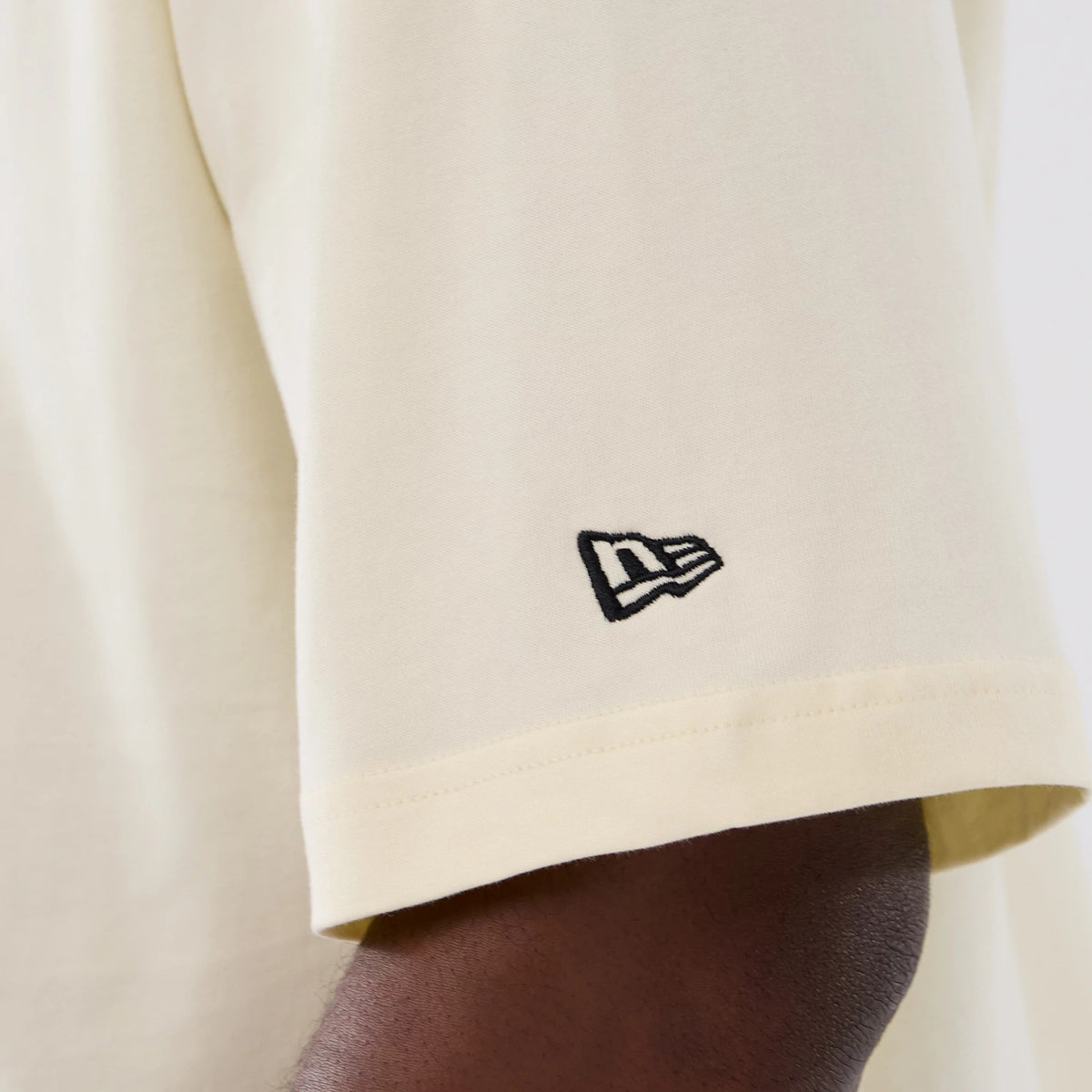 New Era Denver Nuggets NBA Hardwood Classic Cream T-Shirt, side view showing a close-up of the sleeve with a black embroidered New Era logo near the hem on a plain cream fabric, highlighting the simple and classic design.