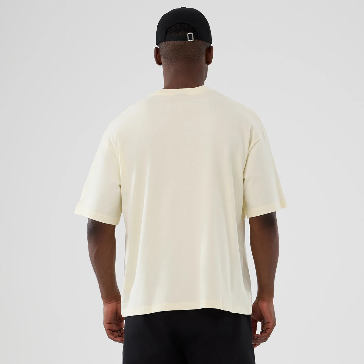 New Era LA Lakers NBA Hardwood Classic Cream T-Shirt, back view showing the plain cream fabric with loose short sleeves and straight hem, worn by a model who pairs it with a black cap.