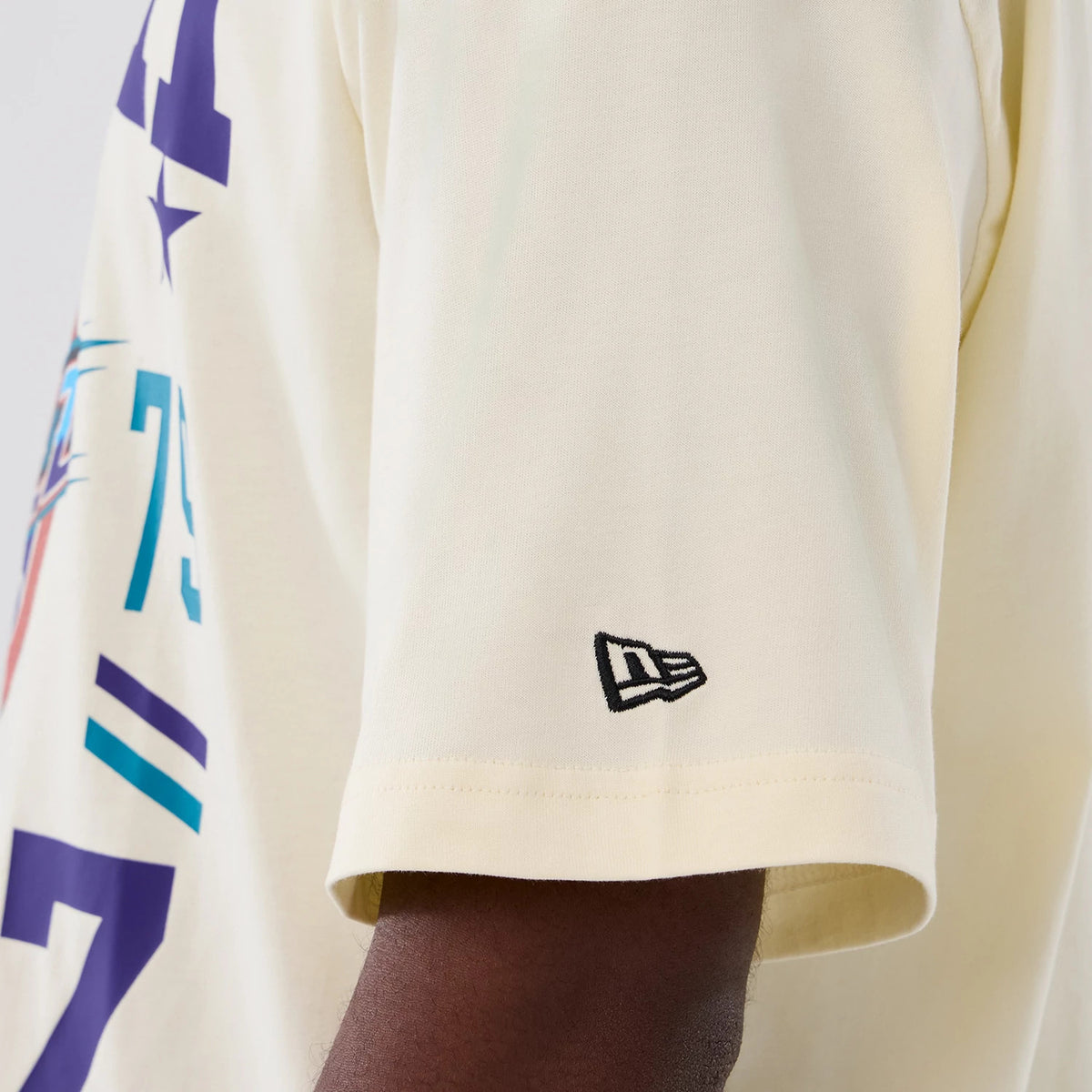 New Era Utah Jazz NBA Hardwood Classic Cream T-Shirt, side view showing a close-up of the sleeve with a small black embroidered New Era logo near the hem and part of the colourful graphic design along the shoulder.