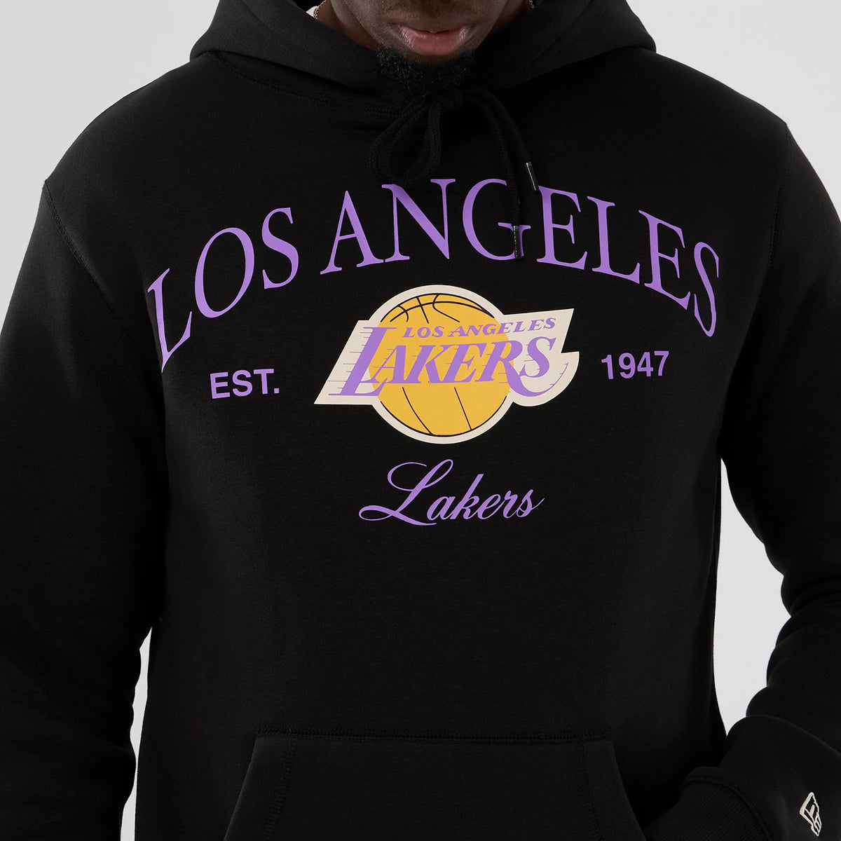 New Era LA Lakers NBA Hardwood Classic Black Pullover Hoodie, front view on-model showing purple Los Angeles Lakers lettering with a yellow basketball graphic on the chest and a small embroidered New Era logo on the sleeve.