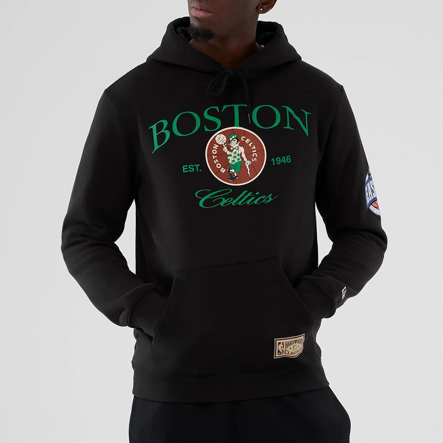 New Era Boston Celtics NBA Hardwood Classic Black Pullover Hoodie, front view on a model showing the green Boston lettering and large Celtics logo on the chest, an embroidered NBA patch on the sleeve and a Classic label above the waistband.