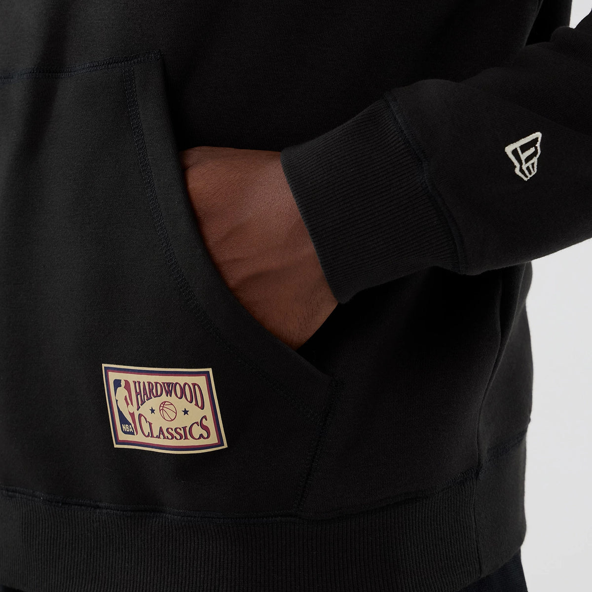 New Era Boston Celtics NBA Hardwood Classic Black Pullover Hoodie, front view showing a hand in the kangaroo pocket with an embroidered Hardwood Classics patch on the pocket, and a small embroidered NBA logo on the sleeve.