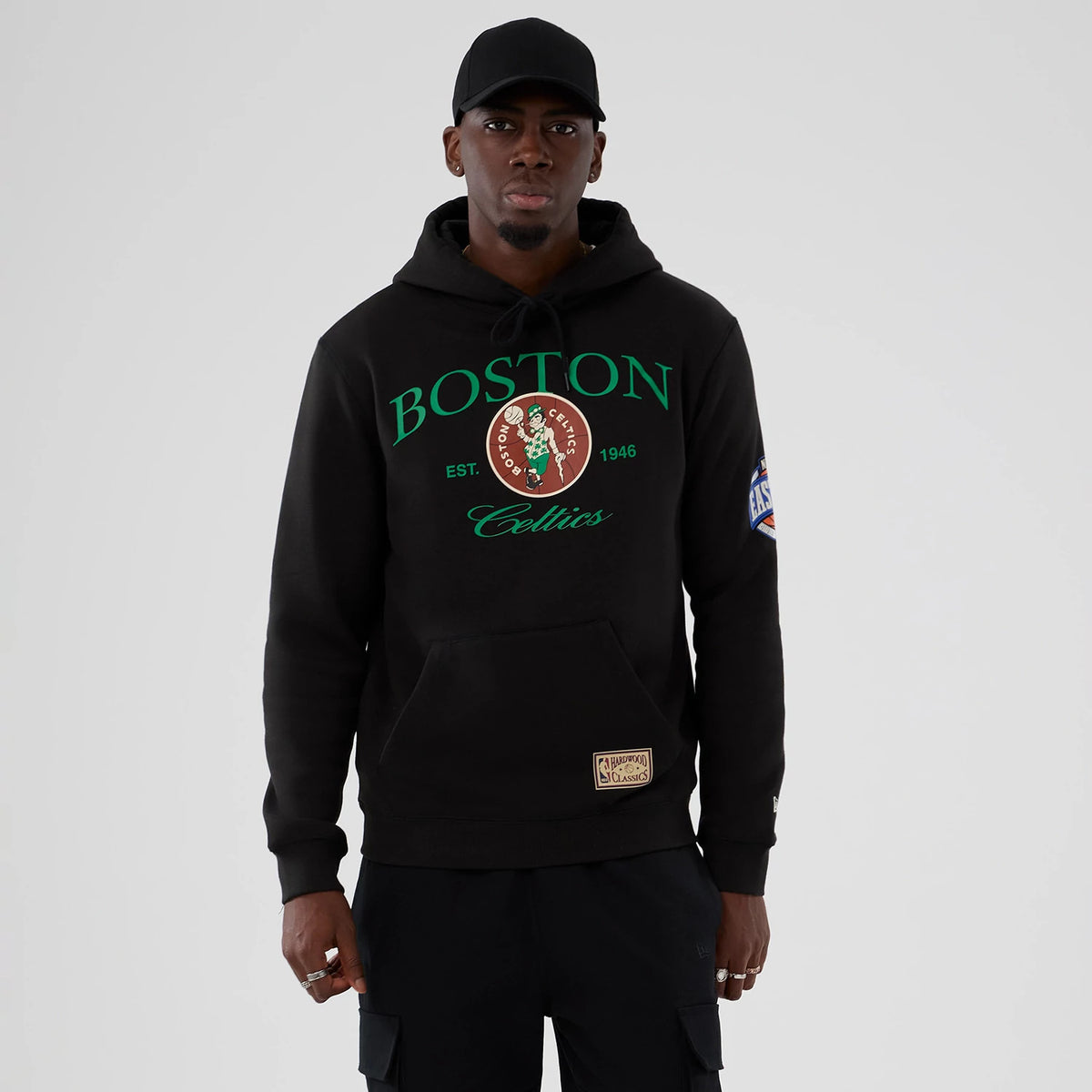 New Era Boston Celtics NBA Hardwood Classic Black Pullover Hoodie, front view on-model showing the green Boston and Celtics text, large Celtics round logo on the chest, and a patch on the lower front, with NBA Hardwood Classics patch on the sleeve.