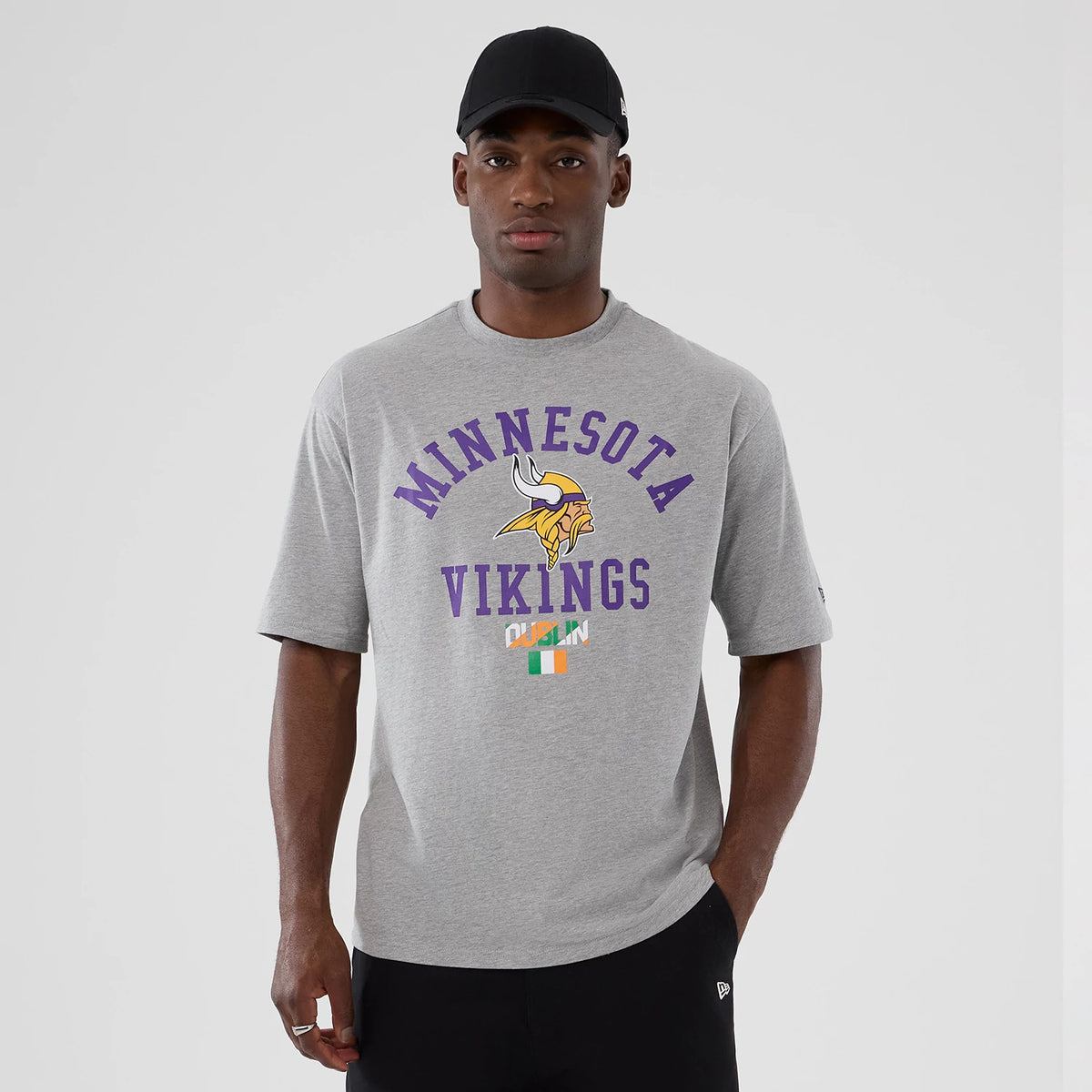 New Era Vikings NFL International Series Games Dublin 2025 Grey Oversized T-Shirt, front view on-model showing the purple Minnesota Vikings lettering and logo on the chest, with an Irish flag graphic below and a printed New Era logo on the sleeve.