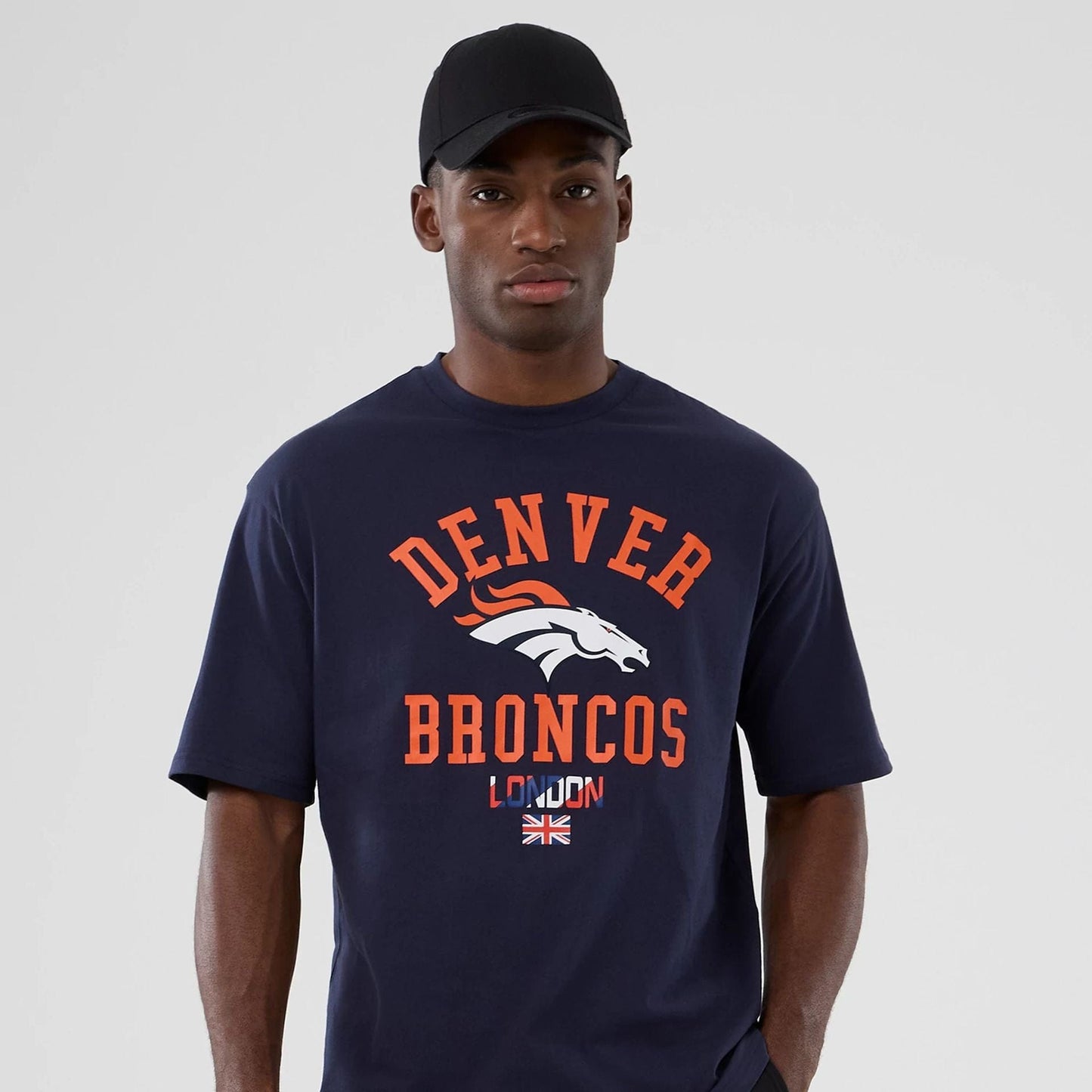 New Era Denver Broncos NFL International Series Games 2025 Navy Oversized T-Shirt, front view on a model wearing a plain black cap, featuring orange Denver Broncos lettering and a white logo above London text with a Union Jack flag graphic.
