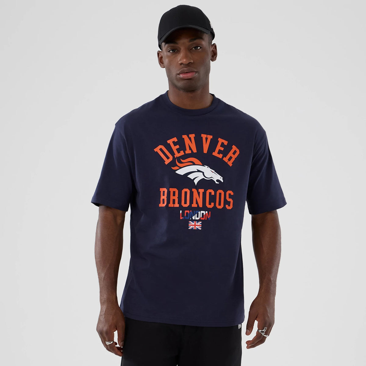 New Era Denver Broncos NFL International Series Games 2025 Navy Oversized T-Shirt, front view on model showing orange Denver Broncos lettering across the chest with a white and orange team logo underneath.