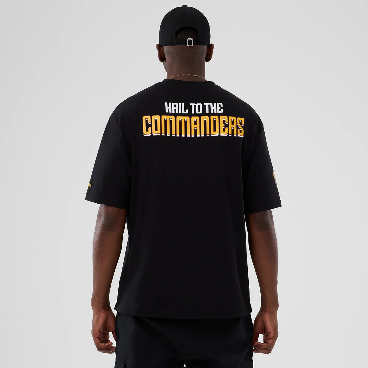 New Era Commanders NFL International Series Games Script 2025 Black Oversized T-Shirt, back view on-model displaying the white and gold embroidered Hail to the Commanders text across the shoulders with a New Era logo on the left sleeve.
