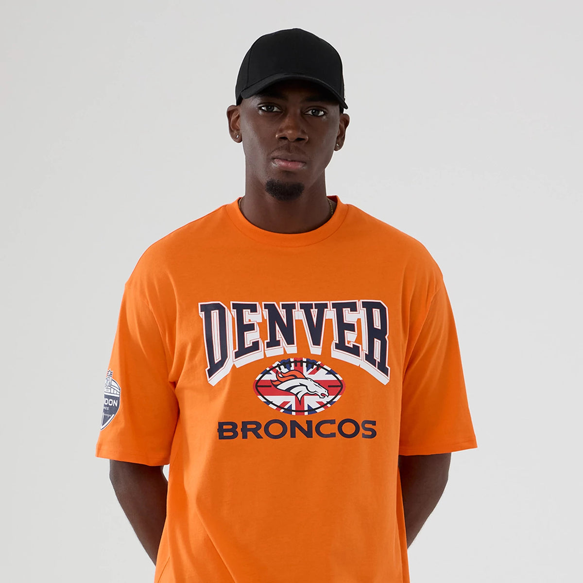 New Era Denver Broncos NFL International Series Games 2025 Orange Oversized T-Shirt, front view on a model with a plain cap, featuring large printed Denver Broncos text and logo on the chest and a commemorative patch on the sleeve.