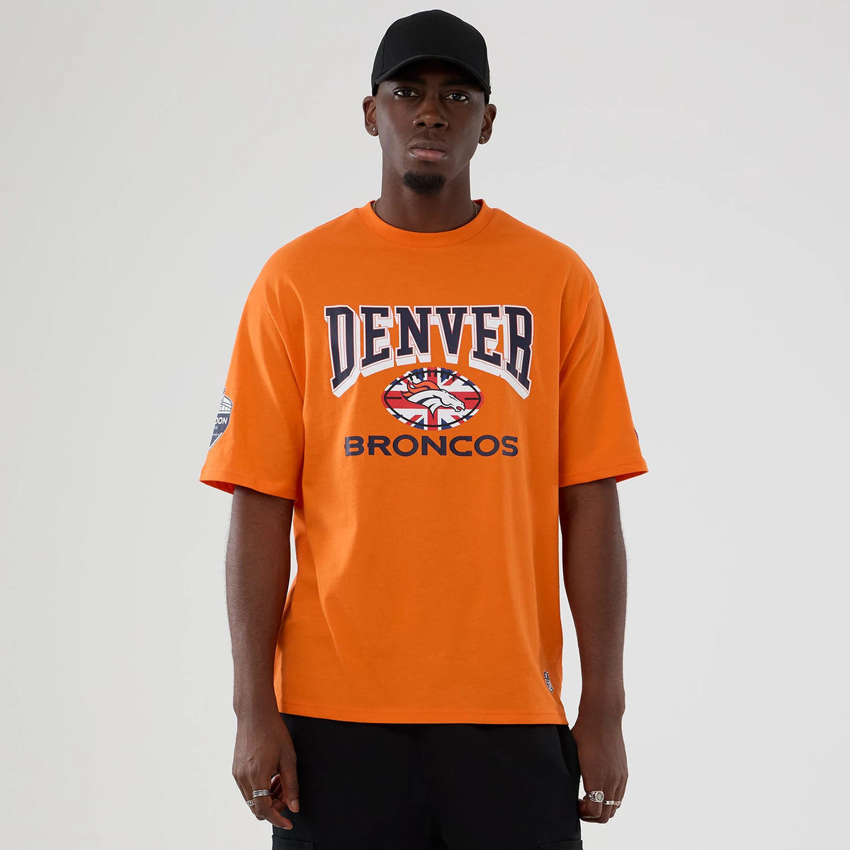 New Era Denver Broncos NFL International Series Games 2025 Orange Oversized T-Shirt, front view on a model showing large embroidered Denver Broncos graphics centred on the chest and a patch on the left sleeve.