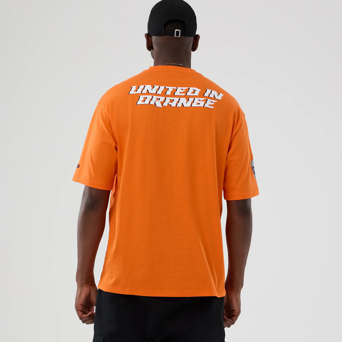 New Era Denver Broncos NFL International Series Games 2025 Orange Oversized T-Shirt, back view on-model featuring white United in Orange lettering across the upper back and a small embroidered New Era logo on the right sleeve.