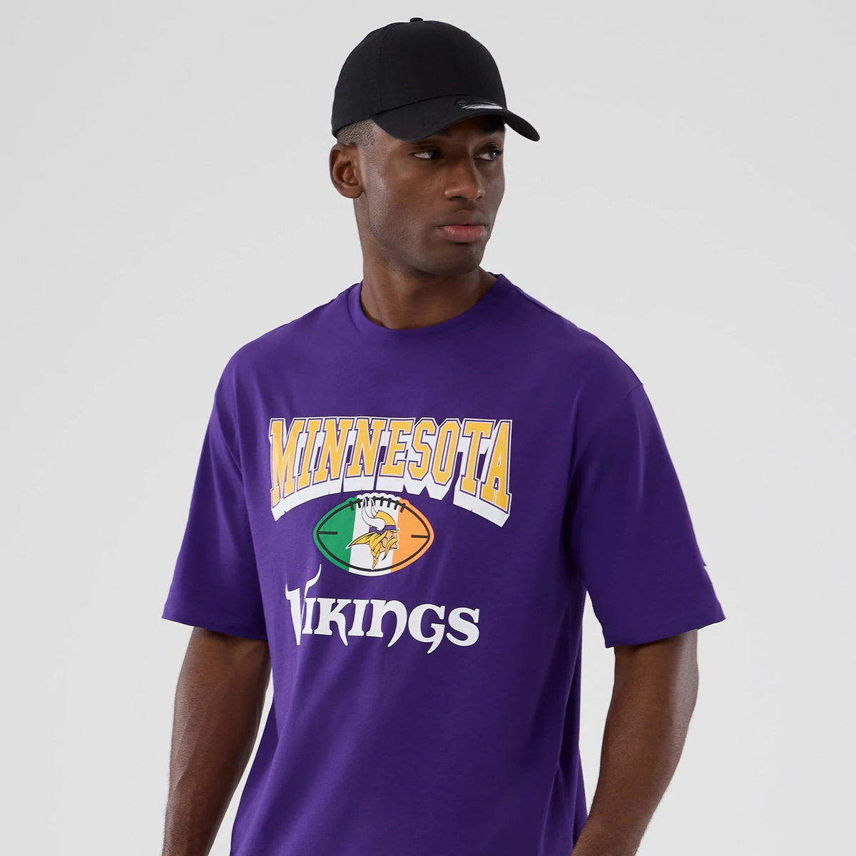 New Era Minnesota Vikings NFL International Series Games 2025 Purple Oversized T-Shirt, front view on-model showing the Minnesota Vikings graphic across the chest with yellow and white lettering against the purple fabric.