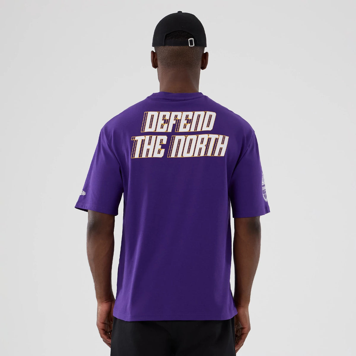 New Era Minnesota Vikings NFL International Series Games 2025 Purple Oversized T-Shirt, back view on a model showing Defend The North text across the shoulders and an embroidered New Era logo on the right sleeve.