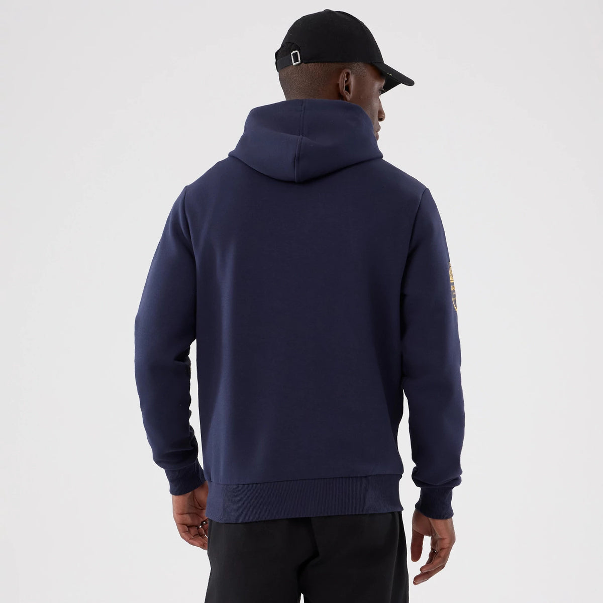 New Era LA Rams NFL International Series Games 2025 Navy Pullover Hoodie, back view on a model wearing a black cap, showing the plain navy rear with a roomy hood and long sleeves featuring a team graphic on the upper left arm.