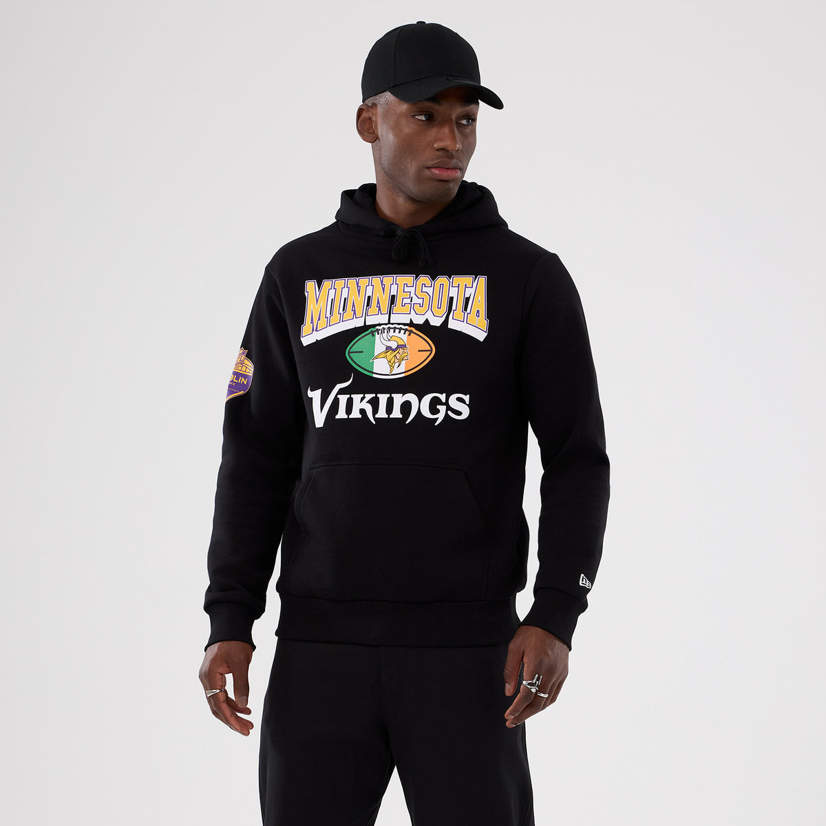 New Era Minnesota Vikings NFL International Series Games 2025 Black Pullover Hoodie, front view on a model showing the large colourful printed logo on the chest, with a small embroidered New Era logo on the left sleeve near the cuff.