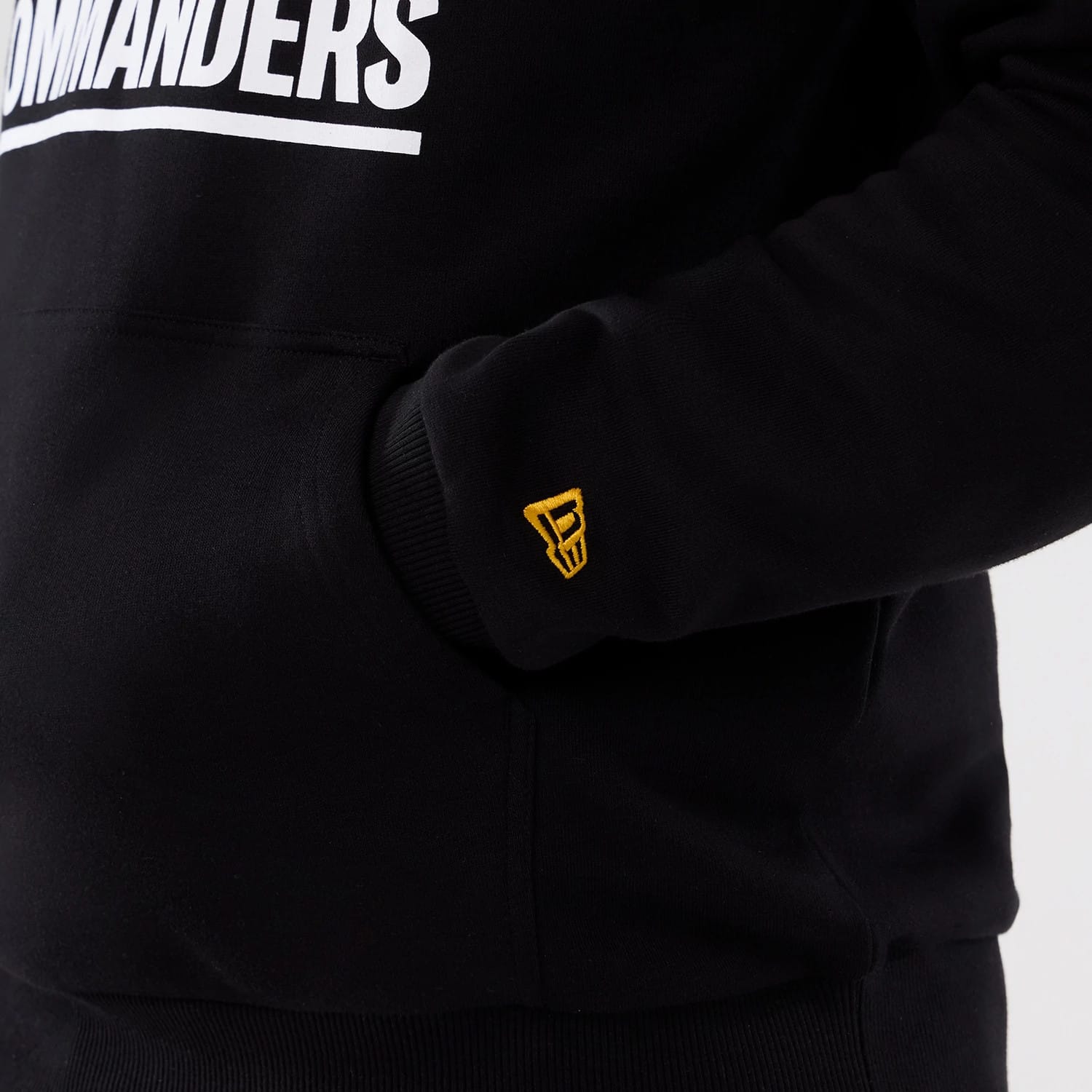 New Era Washington Commanders NFL International Series Games 2025 Black Pullover Hoodie, side view focused on the embroidered gold New Era logo near the cuff, with white Commanders lettering across the chest and the model's hand in the front pocket.