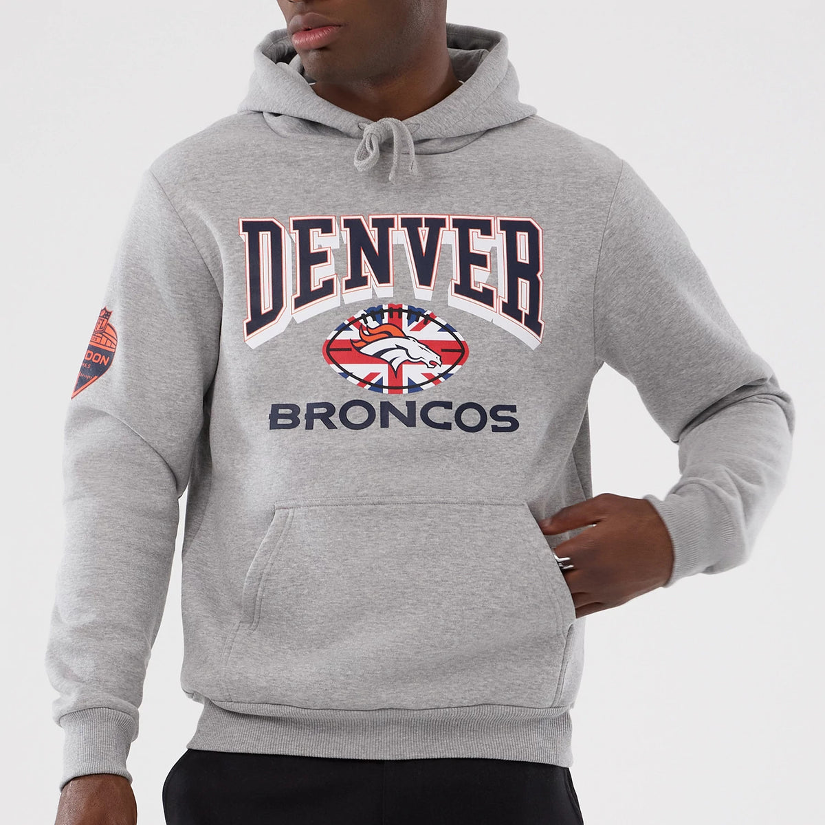 New Era Denver Broncos NFL International Series Games 2025 Grey Pullover Hoodie, front view on-model showing the embroidered Denver letters across the chest, Broncos logo with a Union Jack design below, and an emblem patch on the right sleeve.