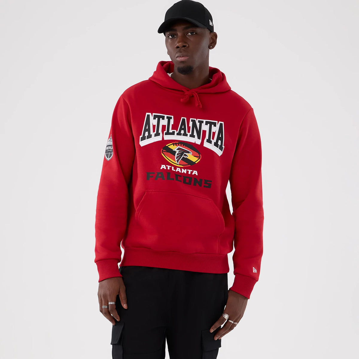 New Era Atlanta Falcons NFL International Series Games 2025 Red Pullover Hoodie, front view on-model showing ATLANTA lettering above the team logo and black Falcons text, with an embroidered New Era logo on the cuff and a patch on the right sleeve.
