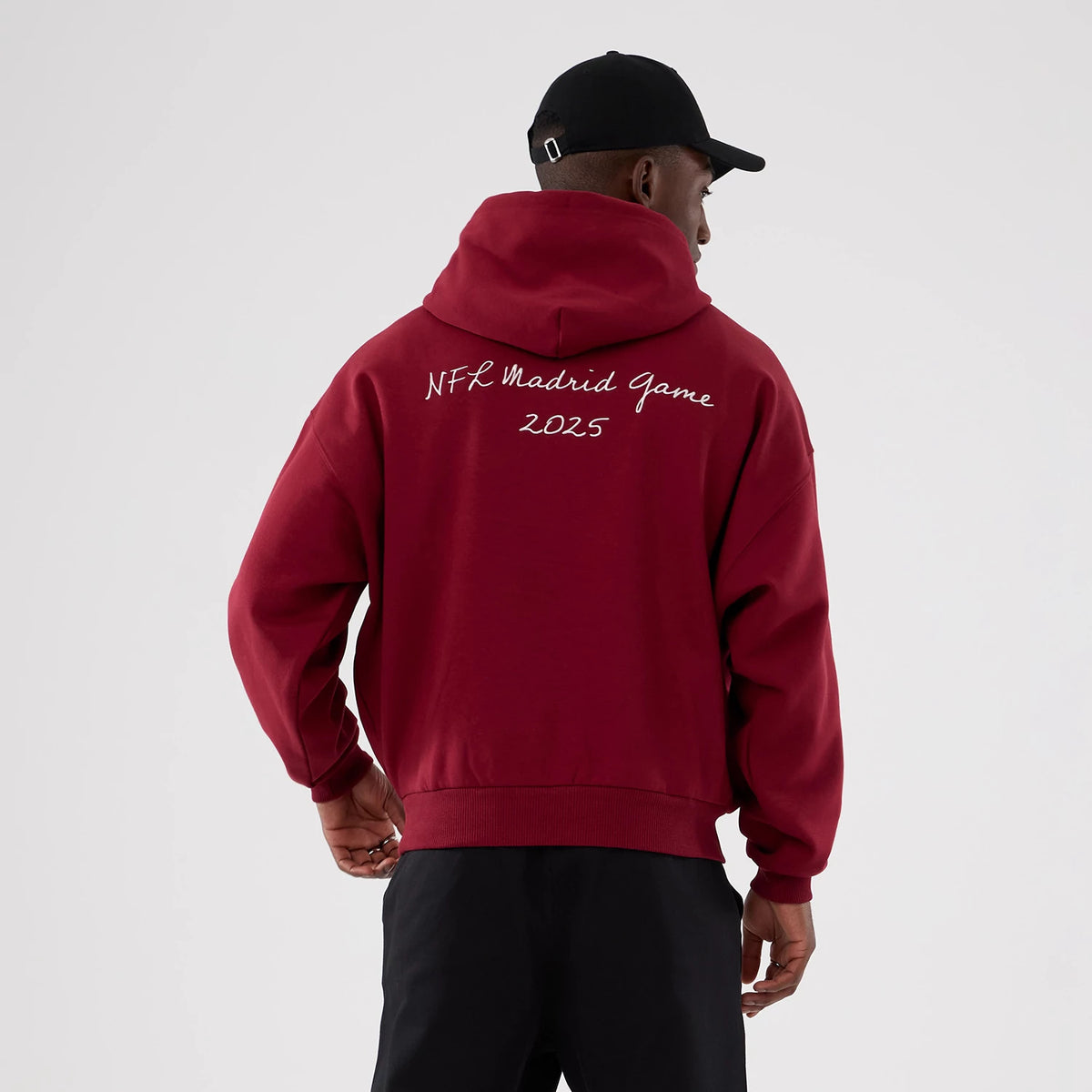 New Era Washington Commanders NFL International Series Games 2025 Dark Red Premium Pullover Hoodie, back view showing the white embroidered NFL Madrid Game 2025 text across the shoulders, styled on a model with dark trousers and a black cap.