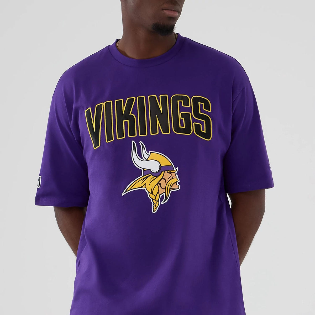 New Era Minnesota Vikings NFL International Series Games 2025 Purple Premium T-Shirt, front view on-model showing the Vikings lettering and Viking head logo printed in gold and white on the chest, with a New Era logo on the left sleeve.