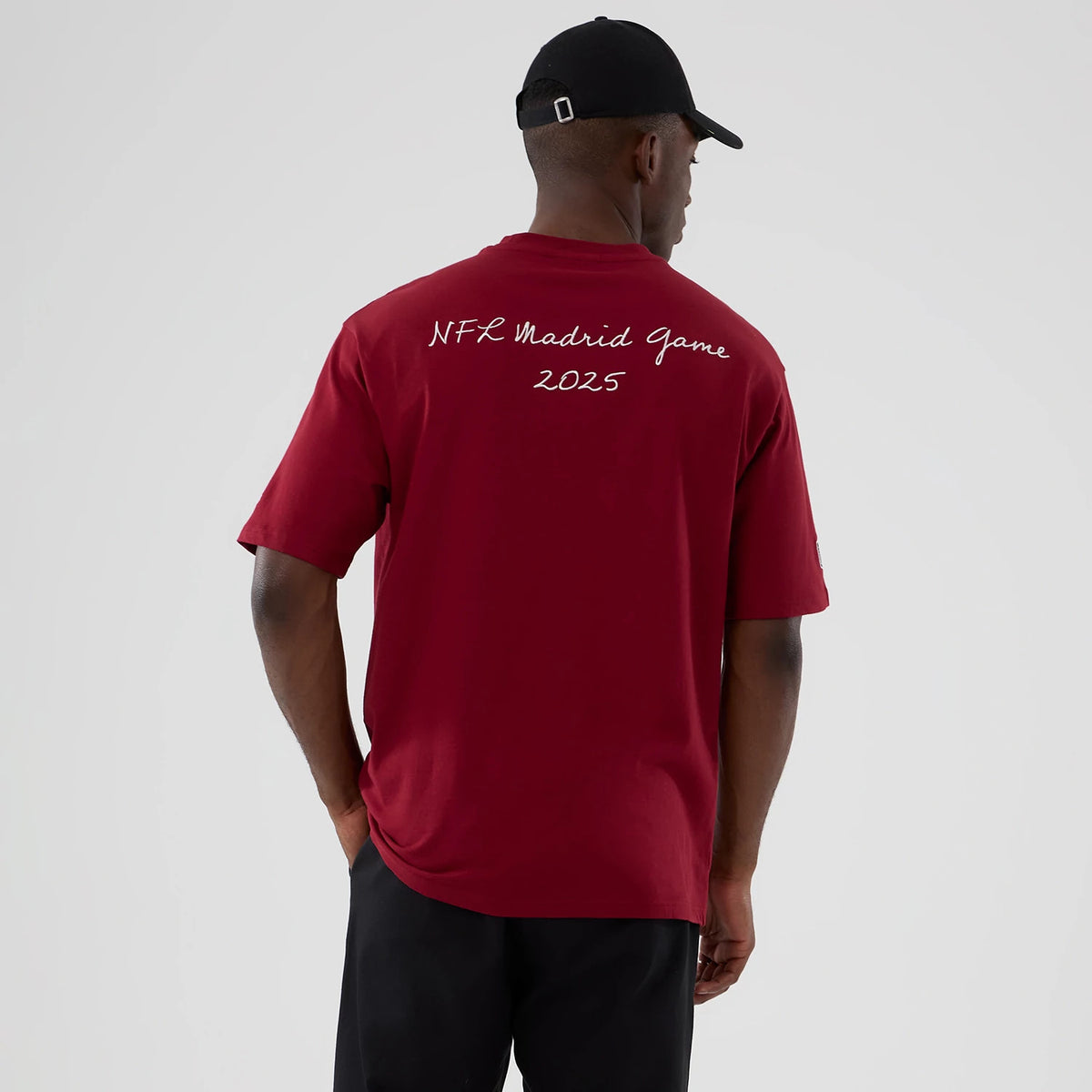 New Era Washington Commanders NFL International Series Games 2025 Dark Red Premium T-Shirt, back view on a model revealing the white embroidered text NFL Madrid Game 2025 centred on the upper back, paired with dark trousers and a black cap.