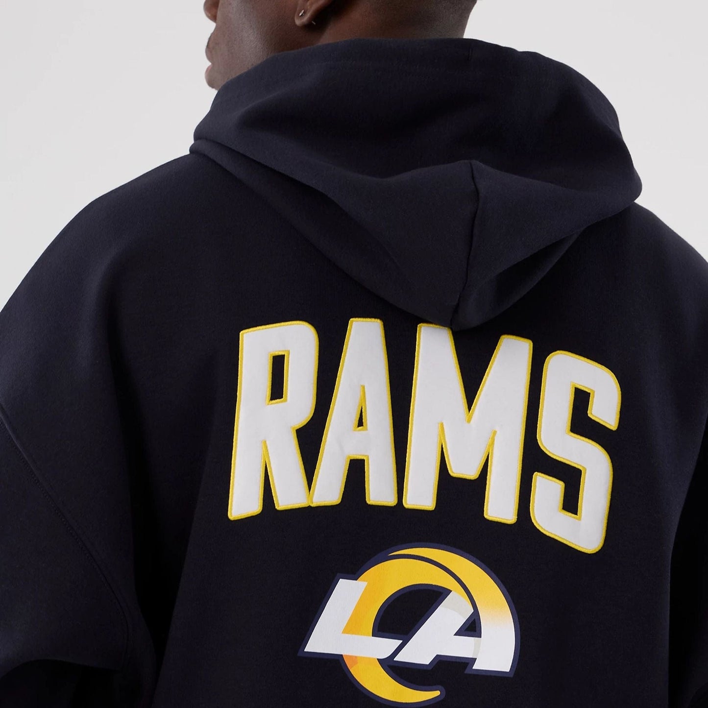 New Era LA Rams NFL International Series Games 2025 Navy Premium Pullover Hoodie, back view on model showing white RAMS lettering outlined in yellow across the upper back and a large printed LA Rams logo underneath on the navy fabric.