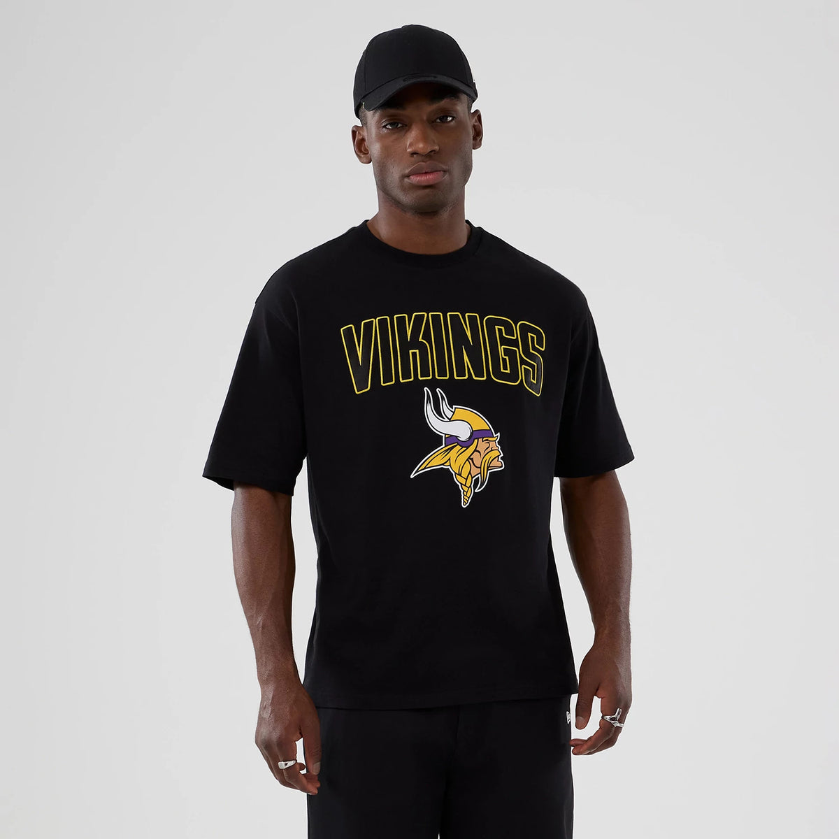 New Era Minnesota Vikings NFL International Series Games 2025 Black Premium T-Shirt, front view on-model showing VIKINGS lettering and a detailed Vikings logo in yellow and white across the chest, paired with a plain black cap.