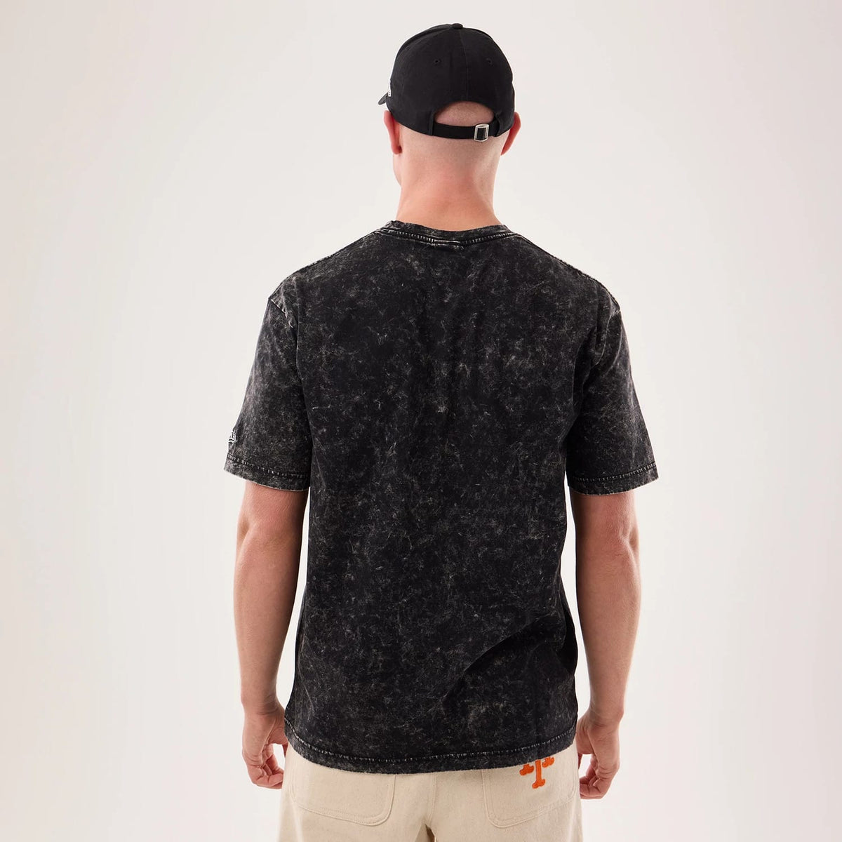 New Era Rick And Morty Science Black T-Shirt, back view on a model showing the textured black fabric with pattern, short sleeves, and a simple crew neckline, paired with light-coloured trousers featuring an orange detail above the pocket.