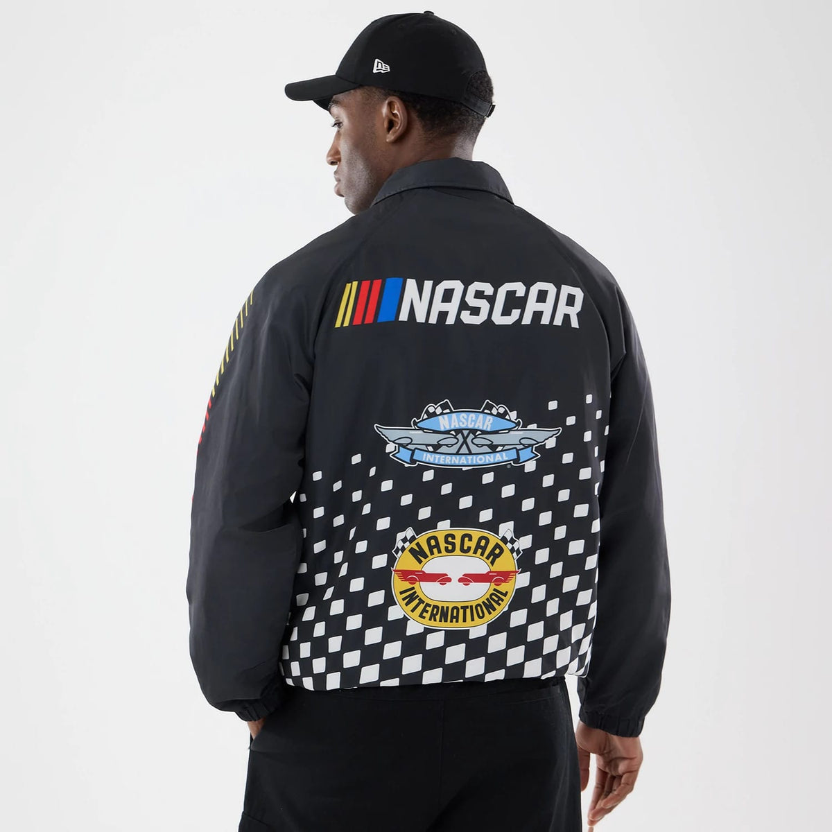 New Era Nascar Logo Black Jacket, back view showing a large printed Nascar logo and graphics in various colours, including checkered patterns at the bottom, worn by a model with a matching black cap and the New Era flag logo on the side.