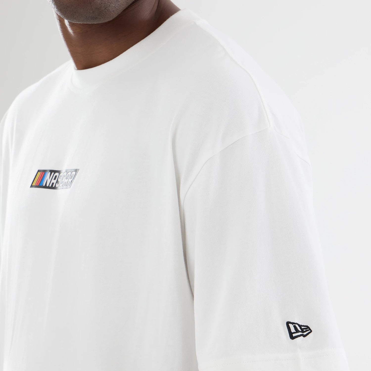 New Era Nascar Logo White T-Shirt, three-quarter view showing the colourful Nascar logo printed on the upper chest, complemented by an embroidered New Era logo on the sleeve, with a classic crewneck and loose fit on-model for casual styling.
