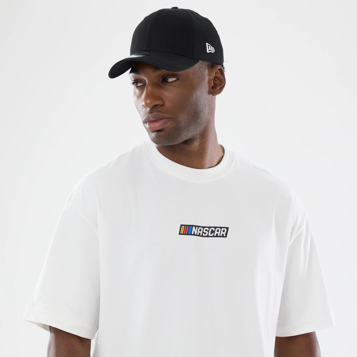 New Era Nascar Logo White T-Shirt, front view on-model featuring the printed Nascar logo centred on the chest in black with coloured stripes, styled with a black New Era cap showcasing an embroidered logo on the left side panel.