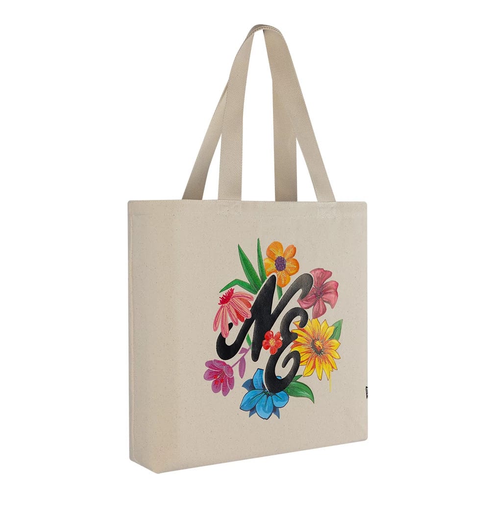 New Era Graphic Light Beige Tote Bag, three-quarter view showing a floral graphic surrounding large stylised black initials on the front, with matching light beige handles extending from the top edges of the bag.