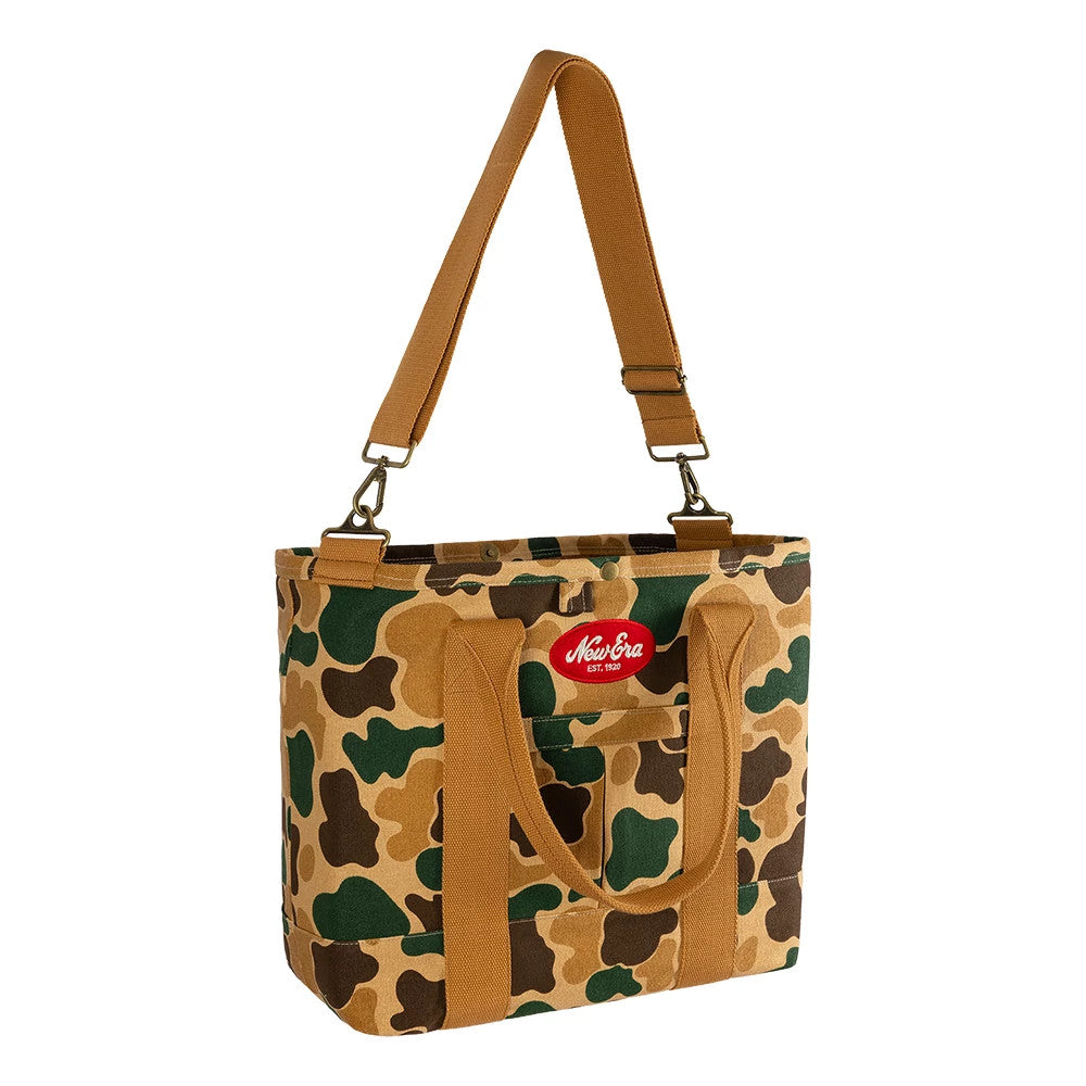 New Era Duck Camo Tote Bag, three-quarter view showcasing the green and brown camo pattern, a red embroidered New Era logo patch on the front, dual brown carry handles, and an adjustable brown shoulder strap attached with clasps.