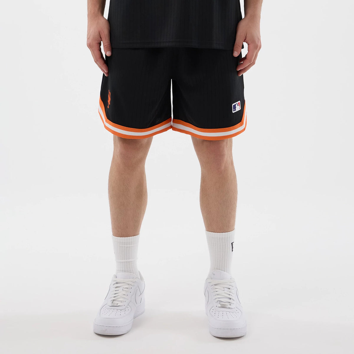 New Era New York Mets MLB Arch Black Oversized Shorts, front view on a model wearing the shorts with double orange stripes along the hem, an embroidered NY logo near the left hem, and an embroidered MLB patch above the right hem.