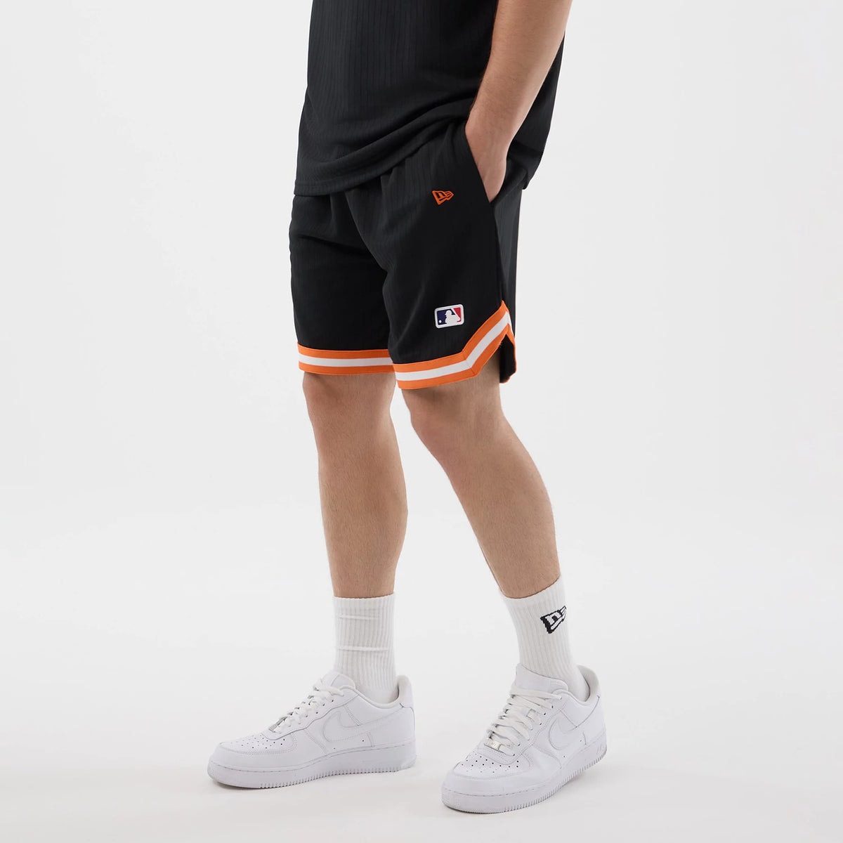 New Era New York Mets MLB Arch Black Oversized Shorts, on-model view displaying the orange and white striped trim on the hems, the embroidered New Era logo on the left leg, and the MLB logo patch near the hem, paired with a black top.
