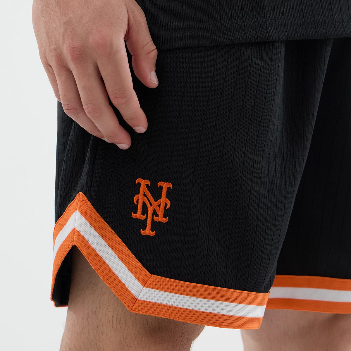 New Era New York Mets MLB Arch Black Oversized Shorts, side view showing the orange embroidered NY logo on the lower leg, distinctive orange and white striped hem, and vertical pinstripes on the fabric, worn on a model.
