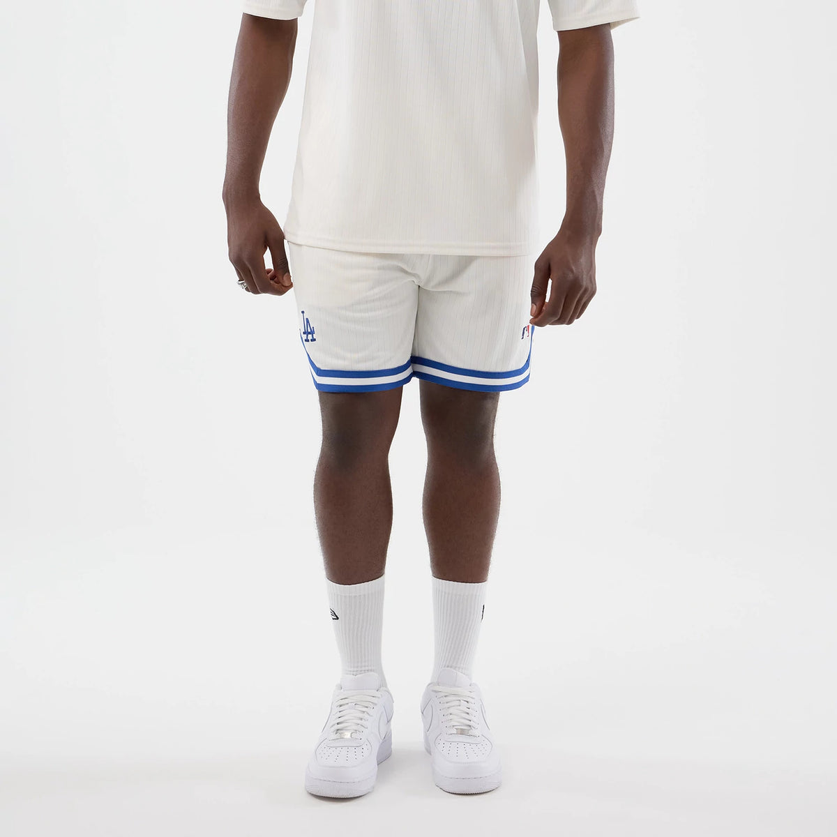 New Era LA Dodgers MLB Arch Open White Oversized Shorts, front view on a model showing the embroidered blue Dodgers logo on the left leg, double blue stripes at the hem, and coordinated styling with a white top and white trainers.