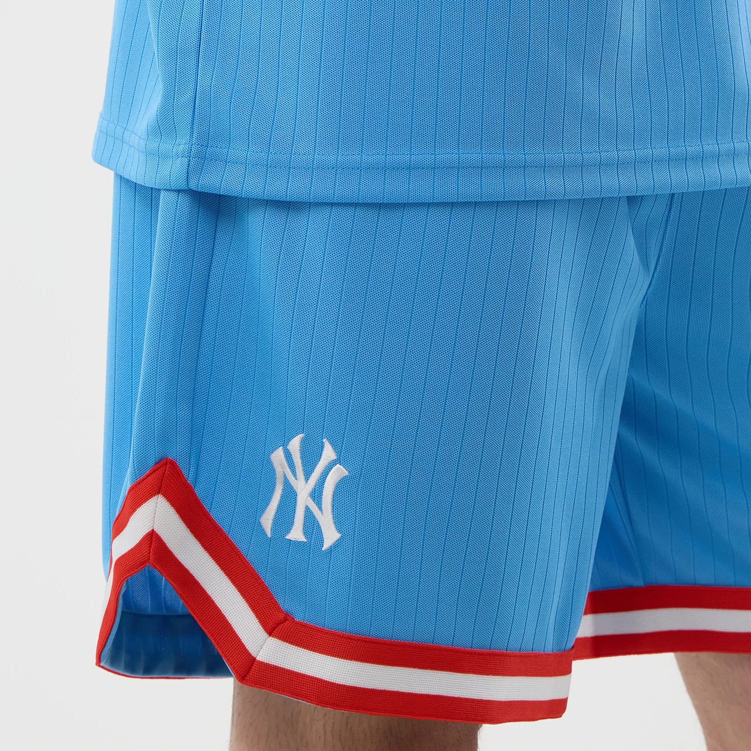 New Era New York Yankees MLB Arch Pastel Blue Oversized Shorts, three-quarter view showing the light blue fabric with vertical pinstripes, the white embroidered Yankees logo on the left leg, and red and white striped trim along the hem.