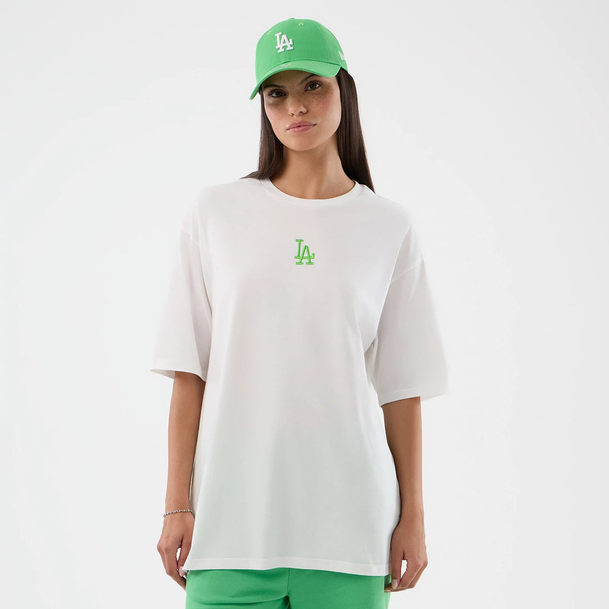 New Era LA Dodgers MLB League Essential White Oversized T-Shirt, front view on a model showing a bright green embroidered LA logo on the chest, the loose-fitting white short sleeves, and styled with a matching green cap and shorts.