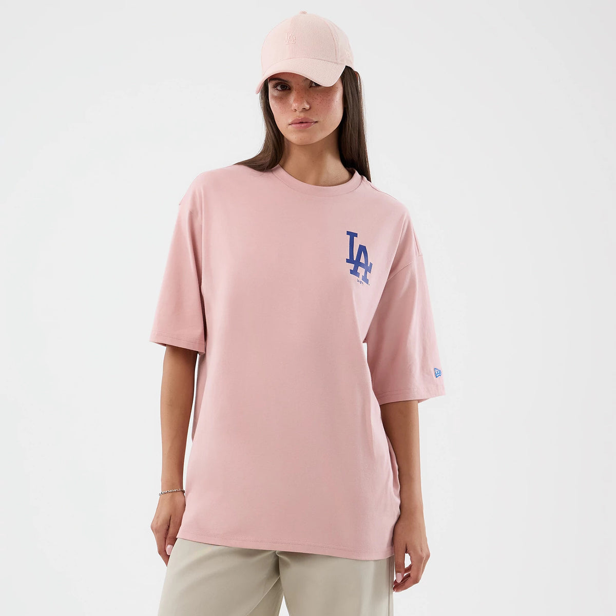 New Era LA Dodgers MLB Cap Graphic Pastel Pink Oversized T-Shirt, front view on-model showing a pale pink T-shirt with a blue printed LA Dodgers logo on the chest and a small New Era logo patch on the sleeve, paired with beige trousers.