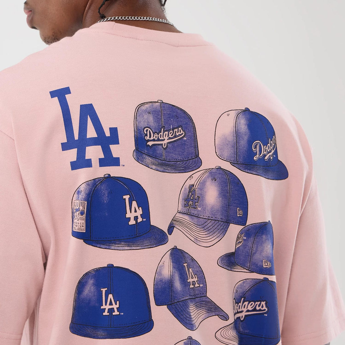 New Era LA Dodgers MLB Cap Graphic Pastel Pink Oversized T-Shirt, back view on a model showing multiple blue cap graphics featuring LA and Dodgers logos, with a large LA logo above, all set against the pastel pink fabric.