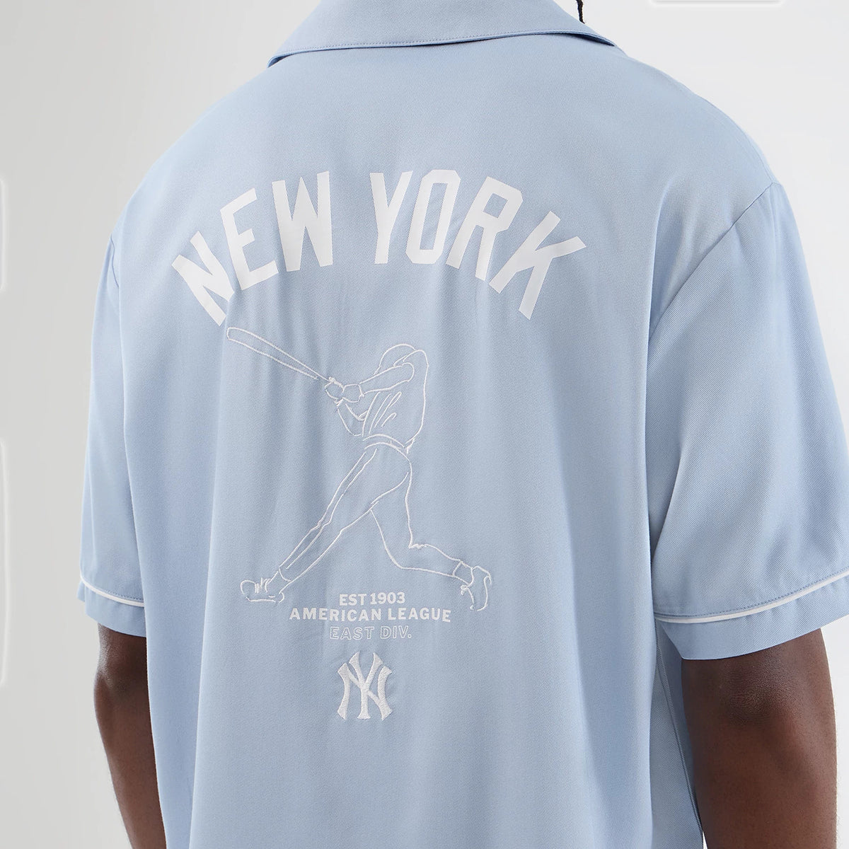 New Era New York Yankees MLB Graphic Revere Pastel Blue Short Sleeve Shirt, back view on a model showing a white New York arch logo and an embroidered outline of a batter swinging a bat, accompanied by the team's logo and established date beneath.