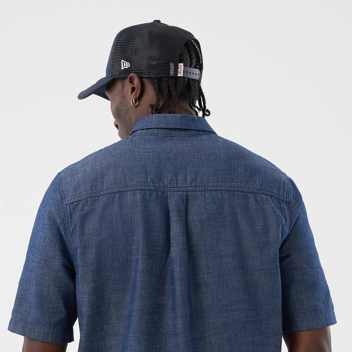 New Era Denim Blue Short Sleeve Shirt, back view on a model wearing a cap, showing the shirt’s classic collar and short sleeves with a straight back yoke, alongside contrast stitching on a textured denim fabric.