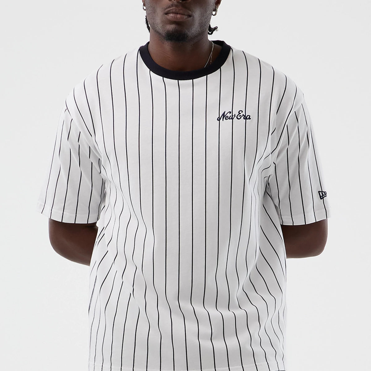New Era Pinstripe Open White Oversized T-Shirt, front view on a model showing vertical black pinstripes, an embroidered New Era logo on the left chest, and a black ribbed collar with a New Era flag logo on the left sleeve.