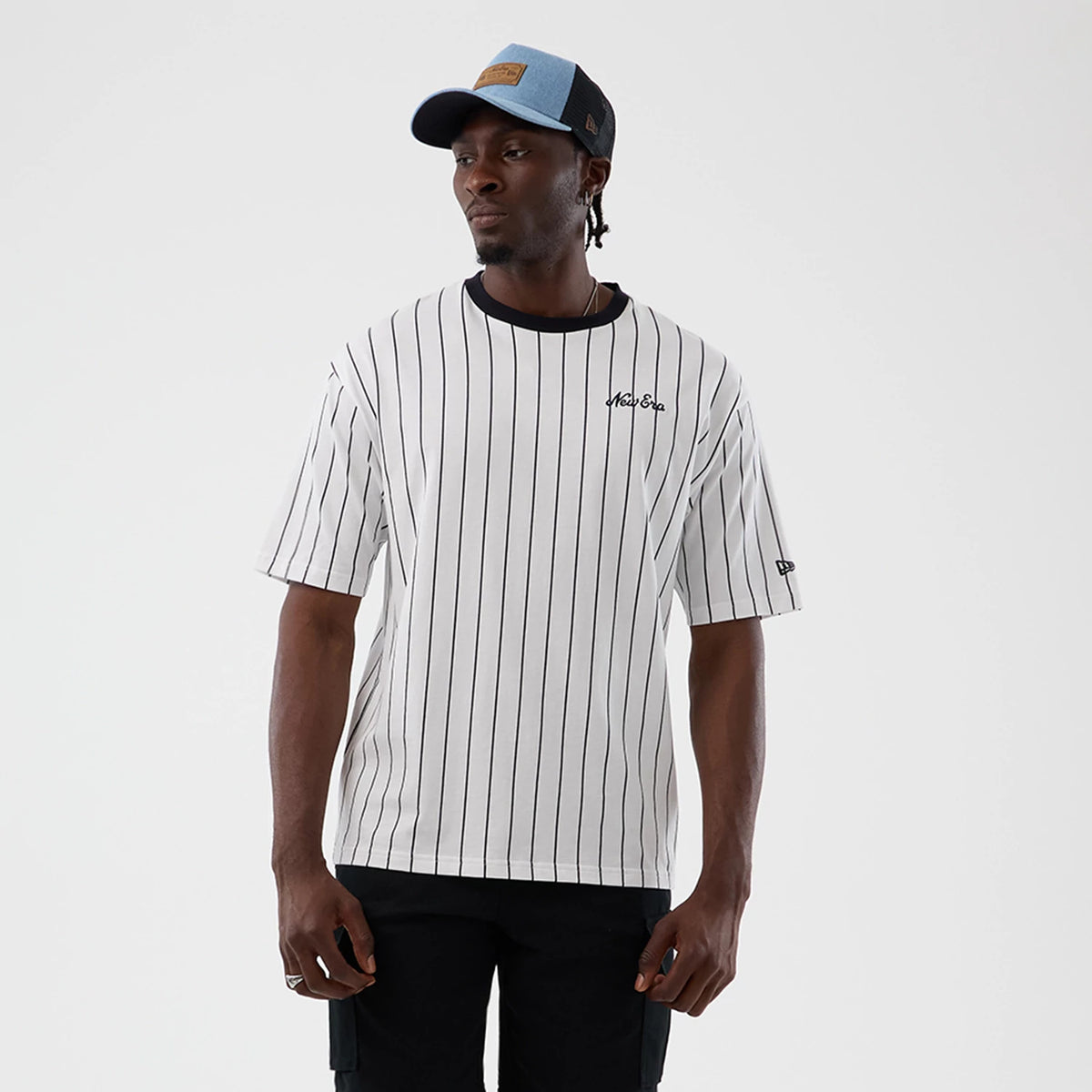 New Era Pinstripe Open White Oversized T-Shirt, front view on a model styled with dark trousers and a cap, showcasing thin black vertical pinstripes, a small embroidered New Era logo on the left chest, and a logo patch on the left sleeve.