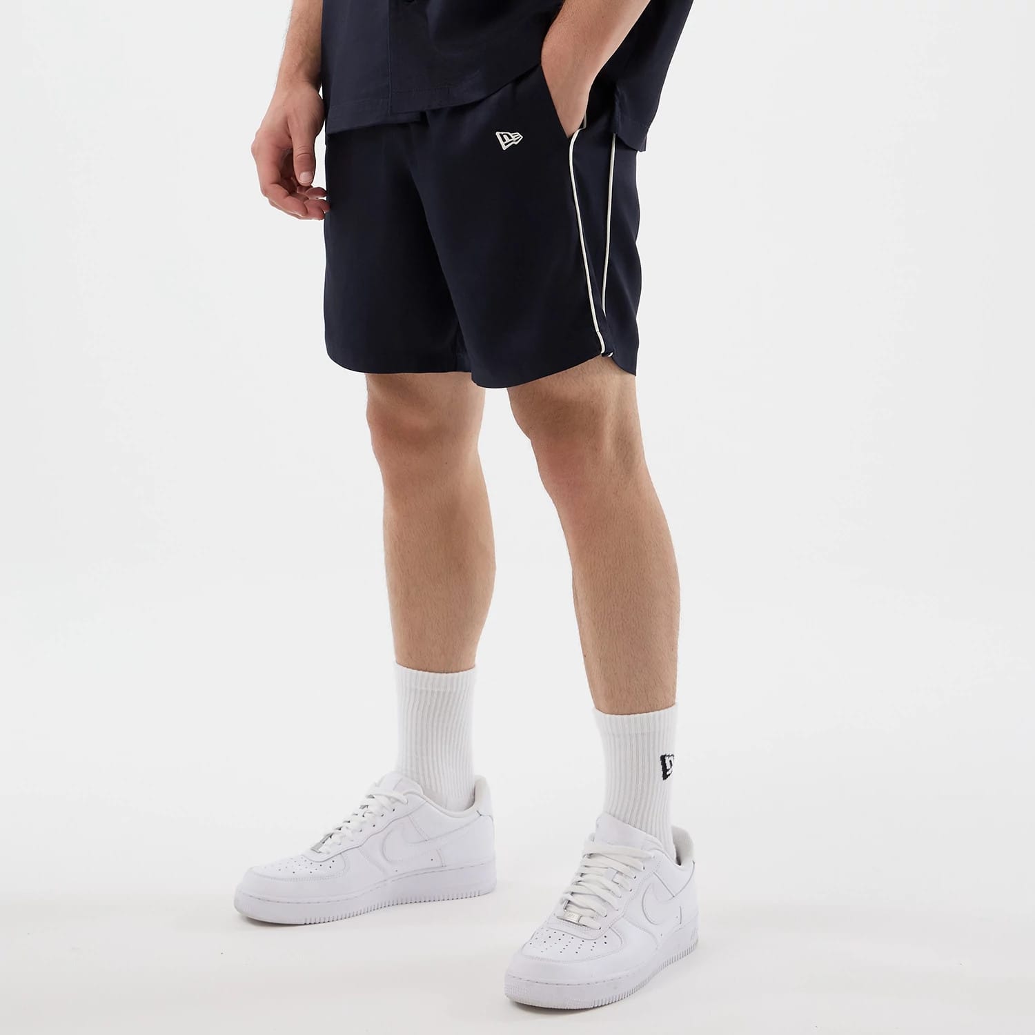 New Era Piping Navy Shorts, on-model view showing the navy shorts with white piping along the side seams, an embroidered New Era logo on the left thigh, with the model wearing white trainers and socks, highlighting the casual styling.