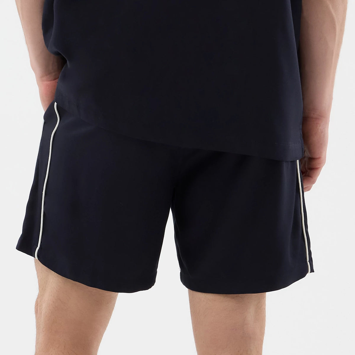 New Era Piping Navy Shorts, rear view on-model showing the dark navy shorts with contrasting white piping running vertically down each side, waistband just beneath a dark top edge, and the model’s hands casually placed near the back seams.
